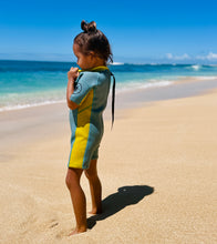 Load image into Gallery viewer, Child in a dusty blue and yellow Selective HA wetsuit standing on a sandy beach with ocean and blue sky in the background